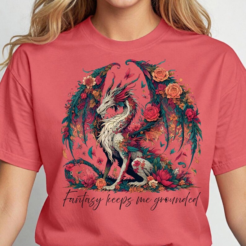 May include: Coral-colored t-shirt featuring a detailed illustration of a dragon with floral wings and accents. The dragon is white with red and green details, surrounded by flowers and butterflies. The text "Fantasy keeps me grounded" is below the dragon.