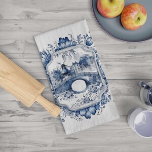 May include: Blue and white kitchen towel with a watercolor design of a windmill and bridge. The design is framed with a floral border.