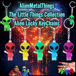 May include: Alien Lucky Keychains in various colors: red, yellow, green, blue, and purple. Each keychain features a small alien figure with outstretched arms, attached to a silver chain and keyring. The text "AlienMetalThings" and "The Little Things Collection" is visible.