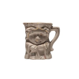 May include: A white ceramic mug with a gold trim and a face sculpted on the front. The mug is shaped like a person and has a handle.