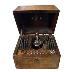 May include: A wooden storage box with a hinged lid containing a collection of metal watchmaker tools. The interior features numerous vertical metal stakes, a central chrome-plated tool, and various small circular metal components arranged in rows.