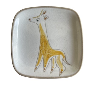 May include: Square ceramic plate with a hand-drawn giraffe illustration. The giraffe is yellow with brown spots and outlines, set against a white background. The plate has a crackled texture and a brown rim, creating a vintage aesthetic.