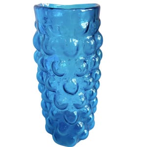 K&ouml;nnte beinhalten: Eine hohe, blaue Glasvase mit einem strukturierten, blasenartigen Design. Die Vase ist aus klarem Glas und hat eine abgerundete, zylindrische Form.