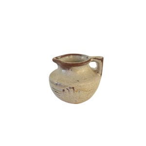 May include: A small, beige ceramic pitcher with a brown rim and handle. The pitcher has a rounded body and a spout for pouring. The surface has subtle, textured markings. A charming, rustic piece for serving.