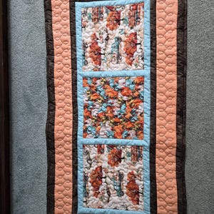 Könnte beinhalten: Gesteppter Tischläufer mit braunem Rand und pfirsichfarbenen Akzenten. Die Mitte zeigt drei rechteckige Paneele mit hellblauem Rand. Die Paneele zeigen ein herbstliches Muster mit Kürbissen und Oldtimern.