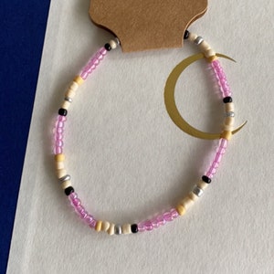 May include: Handmade beaded bracelet with pink, black, cream, silver, and yellow beads. The bracelet is strung on a clear elastic cord. A brown tag with the word "Handmade" and a heart is attached to the bracelet.