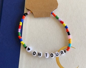 Peut inclure: Un bracelet de perles colorées avec le texte "ON TOUR" en perles de lettres blanches. Le bracelet présente une variété de petites perles rondes multicolores. Une étiquette marron avec le mot "Handmade" et un cœur est attachée.