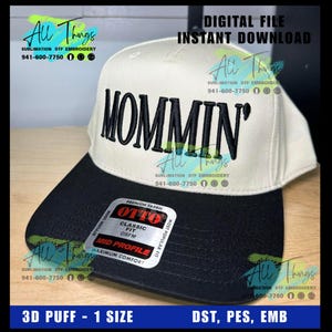May include: A two-tone baseball cap with a black brim and a cream-colored crown. The word "MOMMIN'" is embroidered in bold, black letters across the front. The hat features a "Classic Fit" and "Mid Profile" design.