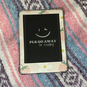 Peut inclure: Une liseuse rose et blanche avec un écran noir affichant un smiley et le texte "PLS GO AWAY I'm reading". La liseuse a un motif de fraises et de fleurs.