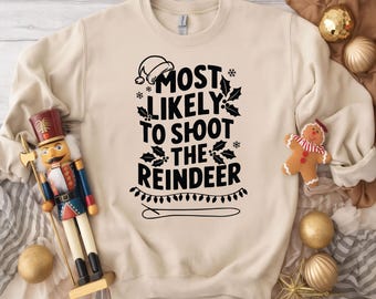 May include: A cream-colored sweatshirt with the text "MOST LIKELY TO SHOOT THE REINDEER" in black. The design includes a Santa hat, snowflakes, holly, and string lights. A wooden nutcracker and Christmas ornaments are also visible.