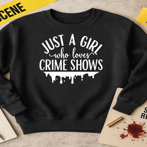 Puede incluir: Sudadera negra con el texto blanco "JUST A GIRL who loves CRIME SHOWS". El diseño incluye un efecto de goteo. La imagen también muestra accesorios de la escena del crimen como cinta de evidencia, un cuchillo y un informe de la escena del crimen.