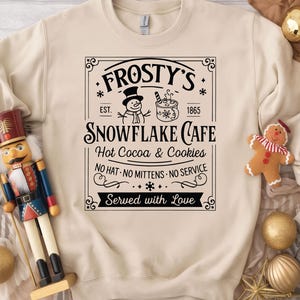 May include: Cream-colored sweatshirt featuring a black graphic for Frosty's Snowflake Cafe, with a snowman, hot cocoa, and the text "Hot Cocoa & Cookies." A wooden nutcracker and Christmas ornaments are also visible.