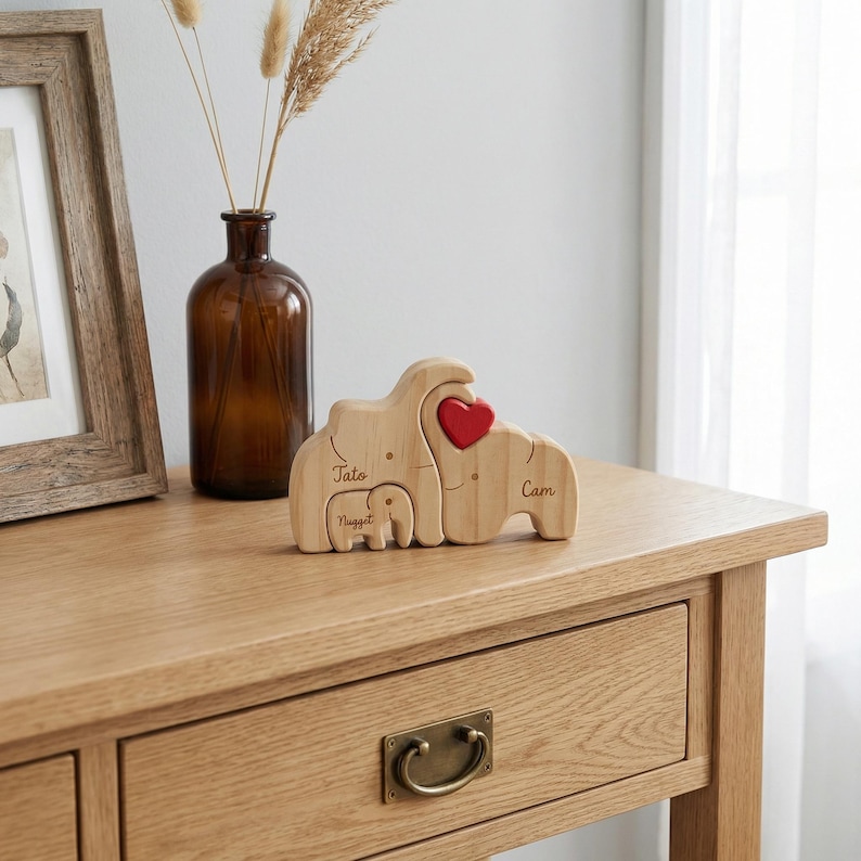 May include: Wooden elephant family figurine with a red heart accent. The names "Jato", "Nugget", and "Cam" are engraved on the elephants. The figurine is displayed on a wooden table with a framed picture and a brown glass vase.
