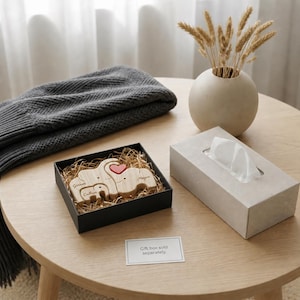 May include: A wooden elephant family set in a black gift box with the names "Garrett", "Megan", and "Callum" engraved. A red heart is placed on the largest elephant. A gray blanket, a vase with dried plants, and a tissue box are also on the table.