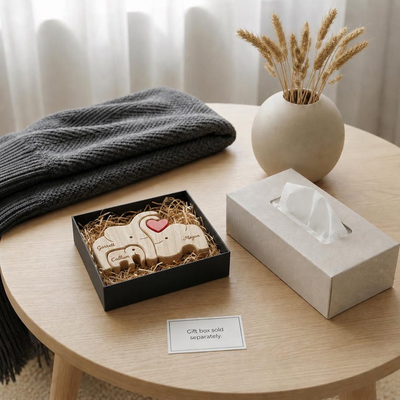 May include: A wooden elephant family set in a black gift box with the names "Garrett", "Megan", and "Callum" engraved. A red heart is placed on the largest elephant. A gray blanket, a vase with dried plants, and a tissue box are also on the table.
