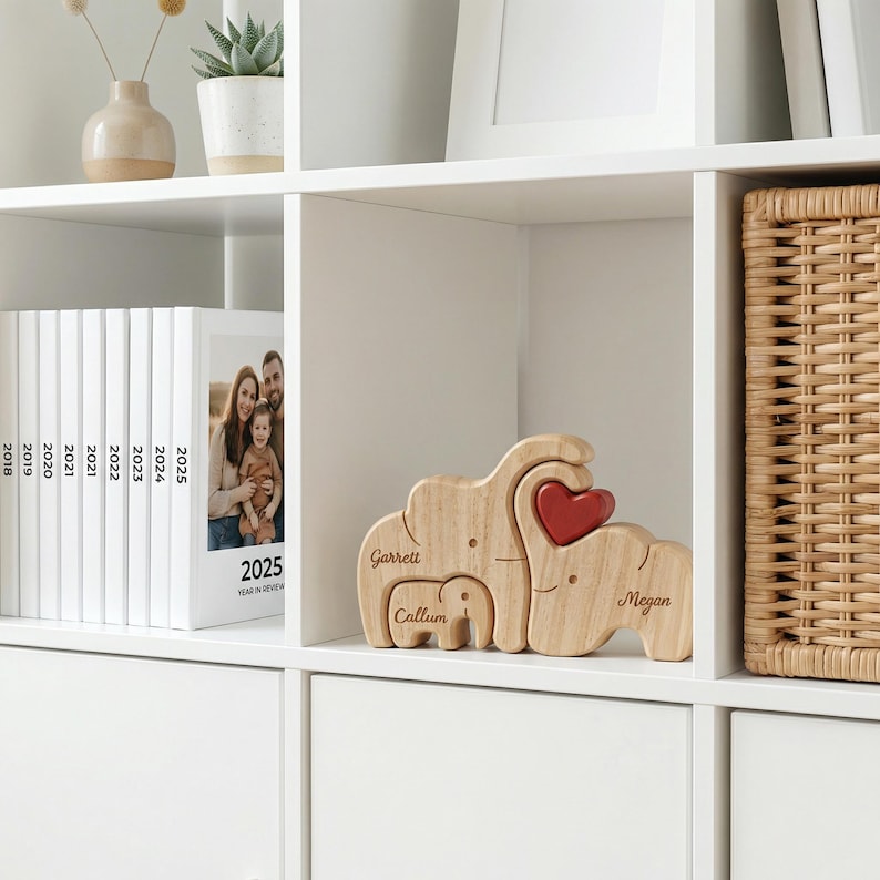 May include: A wooden elephant family puzzle with a red heart detail. The puzzle includes three elephants, each with a name engraved on it. The puzzle is displayed on a white shelf with other decorative items.