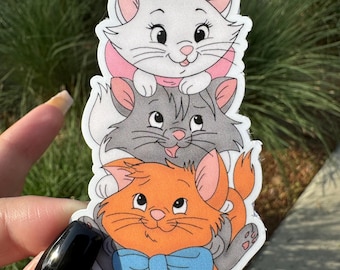 May include: A stack of three cartoon cat stickers. The top cat is white with a pink bow, the middle cat is gray, and the bottom cat is orange with a blue bow tie.