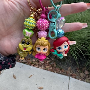 May include: A set of four colorful keychains featuring Disney princess characters. The keychains include a green alligator, a pink princess, a red mermaid, and a blue heart charm. Each keychain has a sparkly bead and a gold or silver ring.