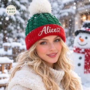 Könnte beinhalten: Eine Wintermütze aus Zopfstrick mit grünem Oberteil, rotem Rippbündchen und einem weißen Bommel. Auf dem roten Bündchen ist der Name Alice in weißer Schrift mit einem kleinen Herzsymbol gestickt. Das Logo Hope Loop ist in der Ecke zu sehen.