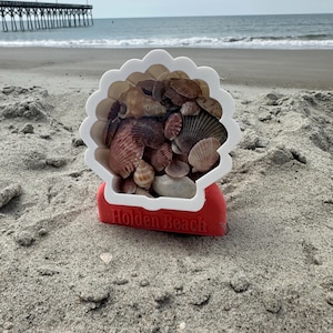May include: A white and red seashell-shaped container filled with various seashells. The container is sitting on a sandy beach with the text "Holden Beach" printed on the base. The ocean and a pier are visible in the background.