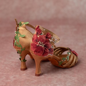 May include: A pair of tan-colored dance shoes with a high heel. The shoes are adorned with red floral embroidery and green leaf accents. The straps are embellished with small, sparkling details. The shoes are designed for ballroom or Latin dance.