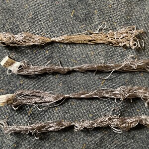 May include: Four bundles of silver and tan embroidery floss are shown against a textured grey background. Each bundle has a small paper tag attached. The floss appears to be made of a shiny, delicate material, ideal for craft projects.