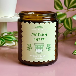 May include: A brown glass jar candle with a label that reads "Matcha Latte." The label has a scalloped edge and features a graphic of a layered drink, leaves, and the text "Soywax Candle 8 oz/250g."