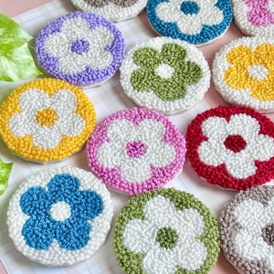 May include: A collection of colorful, handmade coasters with a floral design. Each coaster features a white flower center surrounded by petals in various colors, including yellow, blue, green, pink, and red. The coasters are made with a textured, looped yarn.