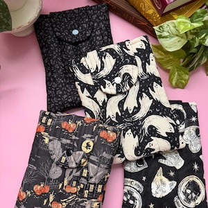 May include: Four rectangular quilted fabric pouches with snap closures. One features a black floral pattern, one has white ghosts on black, one displays an orange pumpkin and house scene, and one shows white skulls and cat heads on a black background.