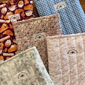 May include: Four rectangular quilted fabric pouches featuring various patterns including a blue gingham check, a dark maroon pastry print, a small floral print, and a white floral line drawing. Each pouch has a white label reading "gloomy little posy".