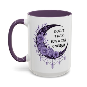 Könnte beinhalten: Weißer Keramikbecher mit lila Innenraum und Henkel. Der Becher zeigt ein Halbmond-Design mit lila Blumen und dem Text "DON'T FUCK WITH MY ENERGY" in schwarzer Schrift. Das Design umfasst hängende Kristallakzente.