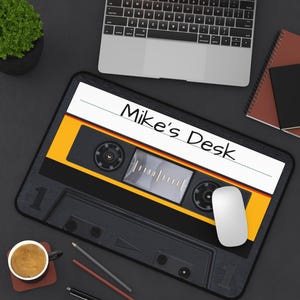 May include: A black and yellow cassette tape-shaped mousepad with the text "Mike's Desk" in white. A white mouse sits on the mousepad. A laptop, notebook, and coffee cup are also on the dark surface.