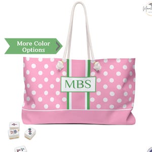 May include: A pink tote bag with white polka dots, featuring a central vertical green stripe and a white rectangular frame containing the green letters MBS. The bag has two white rope handles and is accompanied by four small mahjong tiles.