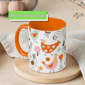 May include: A ceramic mug with a bright orange handle and interior, featuring a white exterior decorated with a repeating pattern of stylized chickens and flowers in shades of orange, pink, and teal, with the text More Colors & Sizes.