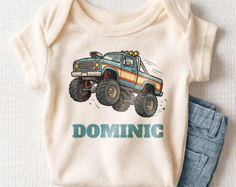 May include: A cream-colored short-sleeve bodysuit featuring a graphic illustration of a blue monster truck with red and yellow stripes. The name DOMINIC is printed in large teal block letters below the truck. A pair of blue corduroy pants.