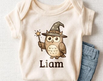 May include: A cream-colored short-sleeve bodysuit featuring a graphic of a brown owl wearing a dark wizard hat with stars, holding a star-tipped wand. The name Liam is printed in bold dark brown letters below the owl illustration.