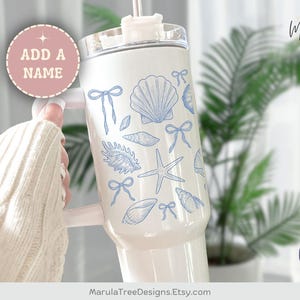 May include: A white tumbler with a handle, adorned with blue seashell and bow illustrations. The text "ADD A NAME" is in a pink circle. The design includes a starfish and various shell shapes. The tumbler is designed and printed in the USA.