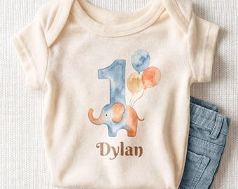 May include: A cream-colored short-sleeve baby bodysuit featuring a watercolor graphic of a blue number one shaped like an elephant, accompanied by three balloons in blue, yellow, and orange. The name Dylan is printed in brown text below.