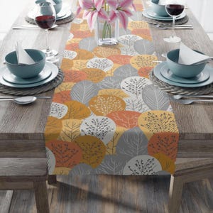 May include: A colorful table runner with a repeating pattern of stylized trees in shades of orange, yellow, and gray. The runner is placed on a wooden table set for a meal, with plates, bowls, and wine glasses.