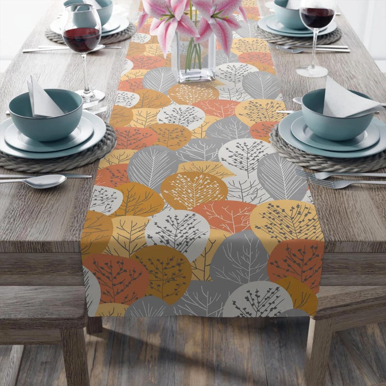 May include: A colorful table runner with a repeating pattern of stylized trees in shades of orange, yellow, and gray. The runner is placed on a wooden table set for a meal, with plates, bowls, and wine glasses.