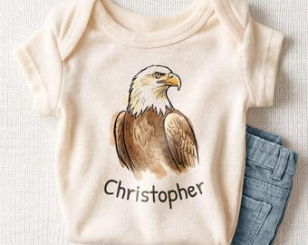 May include: A cream-colored short-sleeve baby bodysuit featuring a watercolor-style illustration of a bald eagle in shades of brown, white, and yellow, with the name Christopher printed in black lettering underneath the eagle graphic.