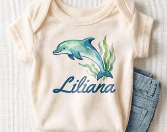 May include: A cream-colored short-sleeve bodysuit featuring a watercolor illustration of a blue and green dolphin swimming among green seaweed, with the name Liliana printed in a dark blue cursive script font below the dolphin.