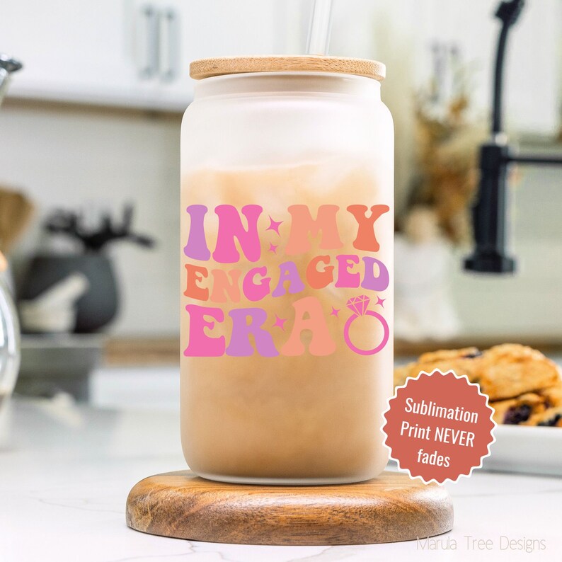 May include: A frosted glass tumbler with a wooden lid and a straw. The tumbler has a pink, orange, and purple graphic that says "In My Engaged Era" with a diamond ring graphic. The tumbler is sitting on a wooden coaster. A red circle with white text says "Sublimation Print NEVER fades".