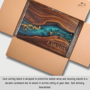 May include: A rectangular wooden cutting board with a turquoise and gold resin design, personalized with the names "Megan & Dave Lawson" and the date "EST. 11.20.2025". The board is inside a cardboard box.