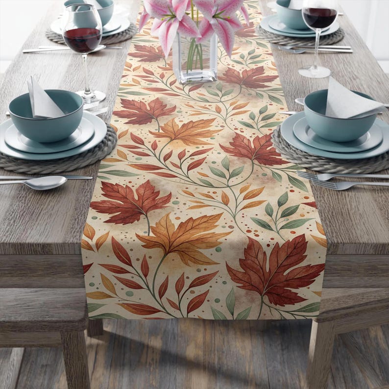 May include: A beige table runner with a repeating pattern of autumn leaves in shades of red, orange, and green. The runner is placed on a wooden table set for a meal, with plates, bowls, and wine glasses.
