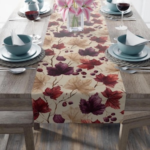 May include: A beige table runner with a repeating pattern of autumn leaves in shades of burgundy, brown, and tan. The runner is placed on a wooden table set for a meal, with plates, bowls, and wine glasses.