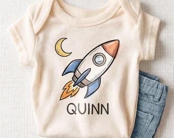 May include: A cream-colored short-sleeve baby bodysuit featuring a graphic illustration of a rocket ship, a crescent moon, and the name QUINN printed in black text, paired with blue corduroy pants.