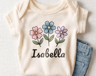 May include: A cream-colored short-sleeve bodysuit featuring a graphic of three flowers in pink, blue, and purple with green stems and leaves. The name Isabella is printed in black script lettering below the floral design, paired with blue denim shorts.