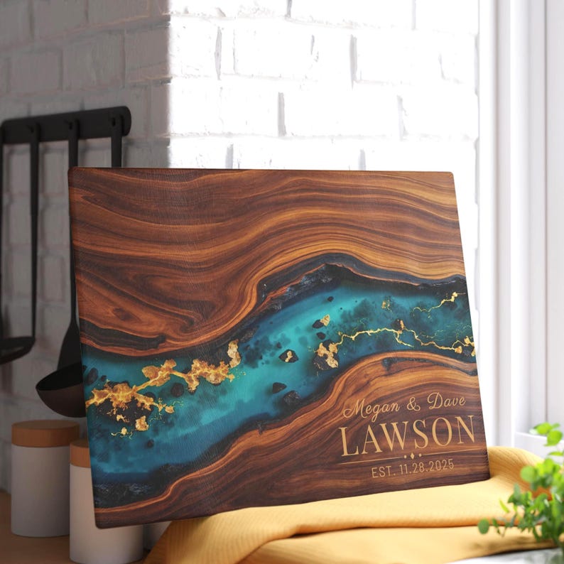 May include: A rectangular wooden cutting board with a turquoise river design and gold accents. The board features the names "Megan & Dave Lawson" and the date "EST. 11.28.2025" in gold lettering. The board is displayed on a yellow cloth.