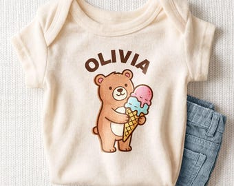 May include: A cream-colored short-sleeve bodysuit featuring a brown bear illustration holding a pink and blue ice cream cone. The name OLIVIA is printed in brown capital letters above the bear. The bodysuit is paired with blue pants.